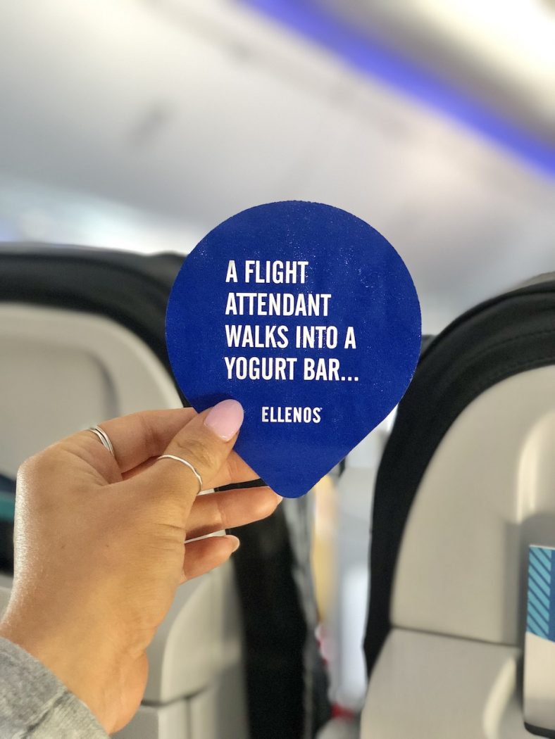 Hand holding a yogurt lid that says, "A Flight Attendant Walks Into a Yogurt Bar... Ellenos" on an airplane.